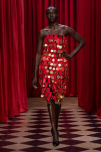 Model in a strapless red dress covered with gold coins on a runway with red curtains behind her.