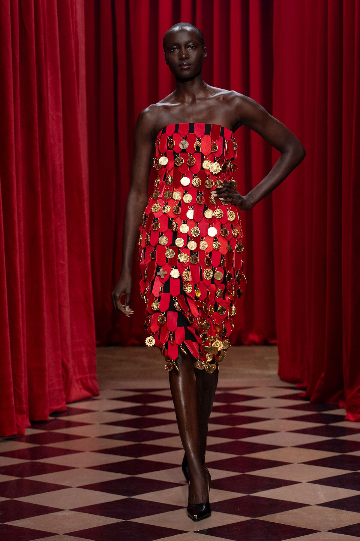 Model in a strapless red dress covered with gold coins on a runway with red curtains behind her.