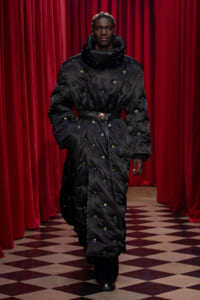 Model wearing a long black quilted puffer coat with gold eyelets and a belt, walking on a checkered floor with red curtains in the background.