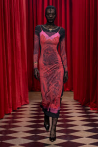 Model walking down a checkered floor in a red dress with black lace sleeves, against red stage curtains, wearing black gloves and heels.