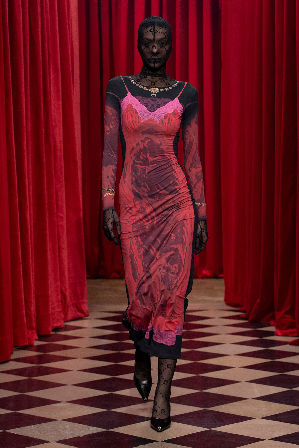Model walking down a checkered floor in a red dress with black lace sleeves, against red stage curtains, wearing black gloves and heels.