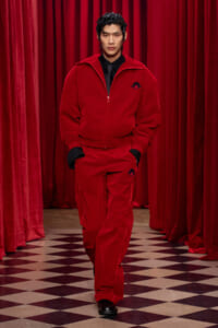 Male model walking the runway in a red tracksuit with a black shirt and tie against red velvet curtains and a black-and-cream checkered floor, hands in pockets.