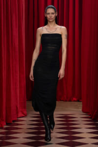 Woman in a black, ruched, strapless dress walking on a red-curtain stage with checkered floor.