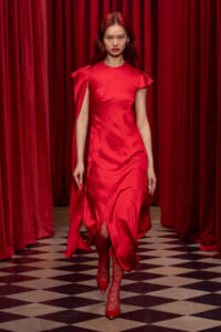 Model walking a runway in a bright red satin dress with a side slit, burgundy curtains in the background.