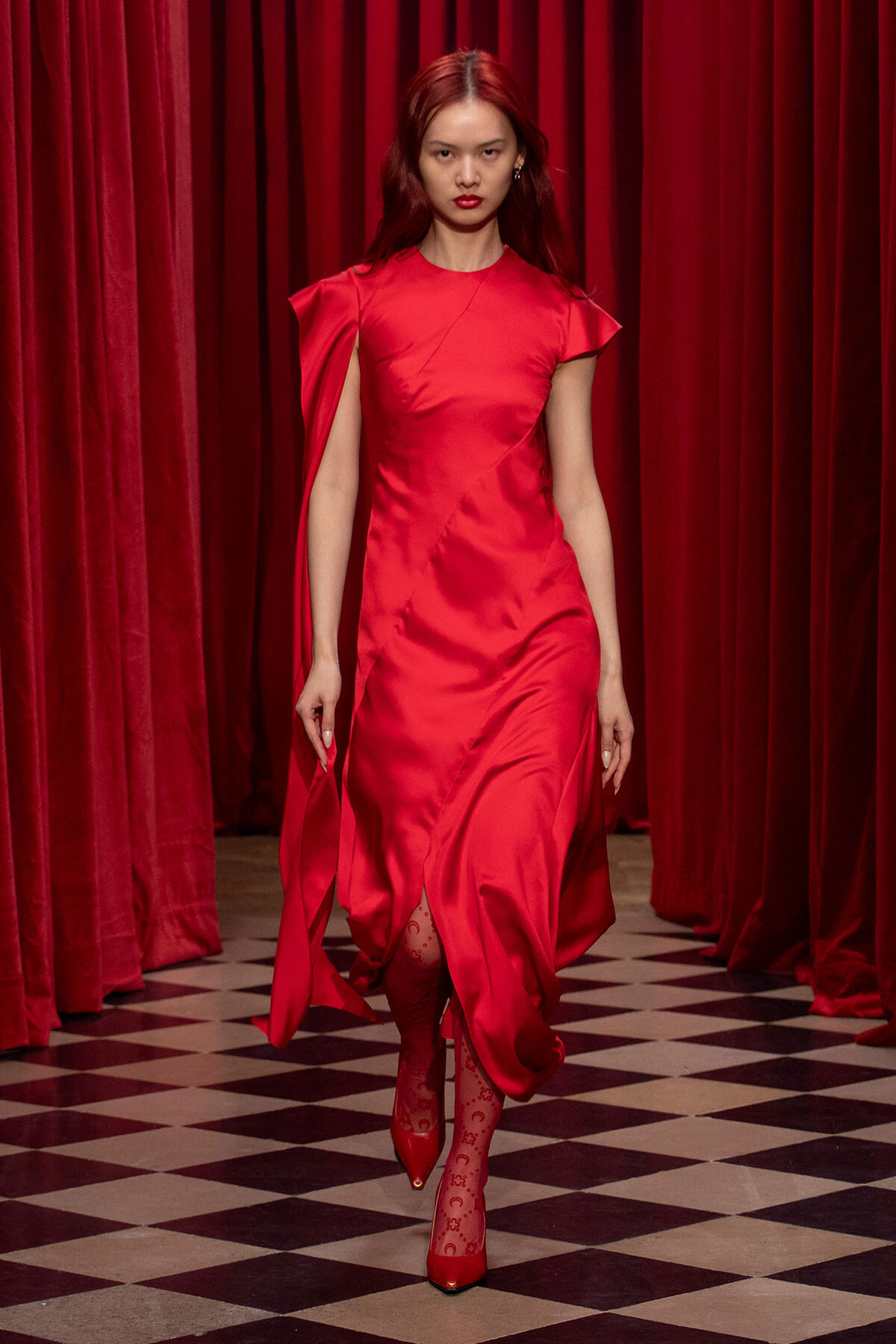 Model walking a runway in a bright red satin dress with a side slit, burgundy curtains in the background.
