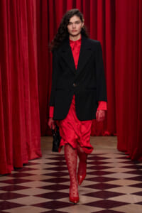 Model walking a runway in a red satin dress under a black blazer, red gloves, and red patterned tights on a checkered floor.