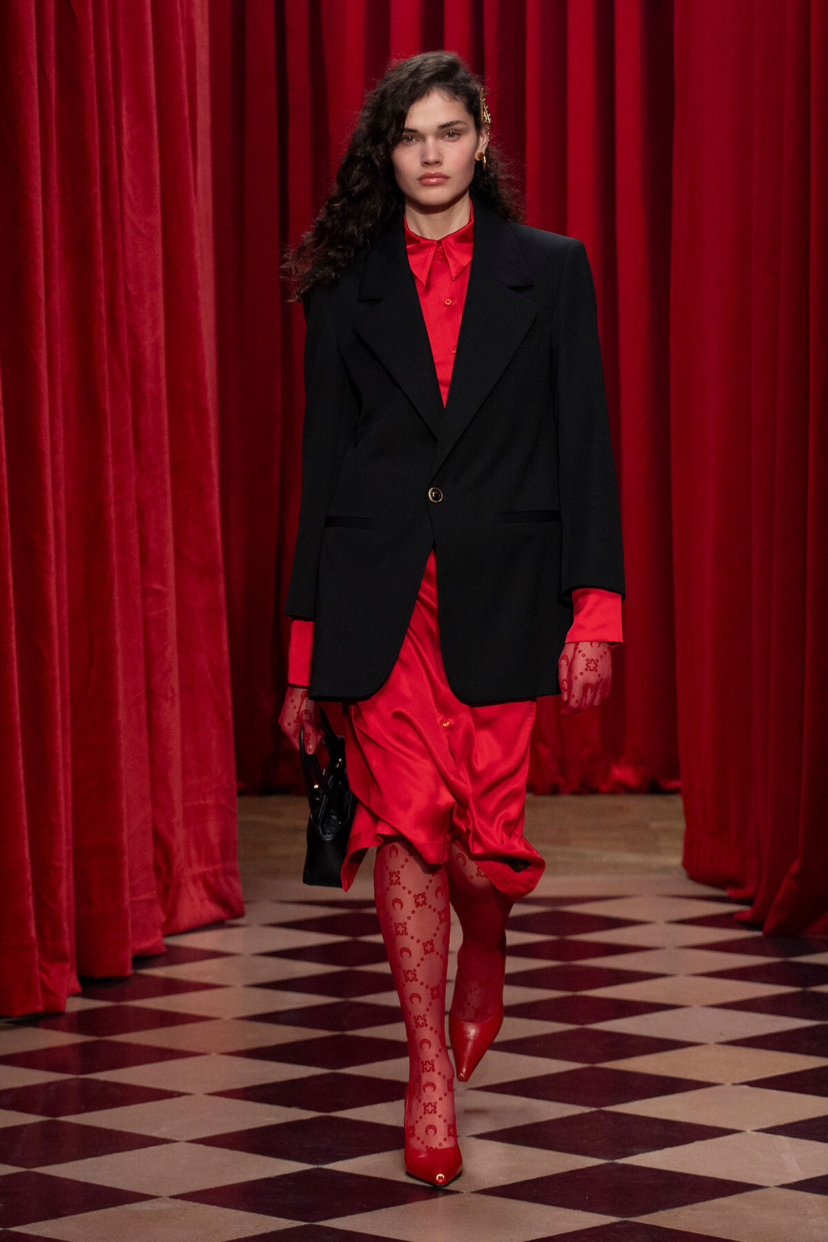 Model walking a runway in a red satin dress under a black blazer, red gloves, and red patterned tights on a checkered floor.