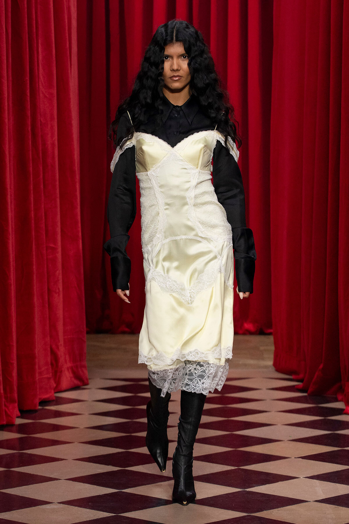 Model wearing a pale cream satin dress with lace overlays over a black blouse, walking down a runway with red curtains behind her.