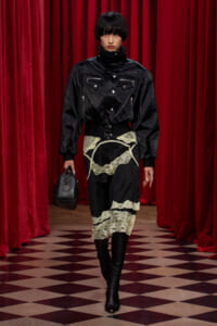 Model in black leather bomber with zip pockets, lace-overlay skirt, and knee-high boots on a red-curtain runway with a small black handbag.