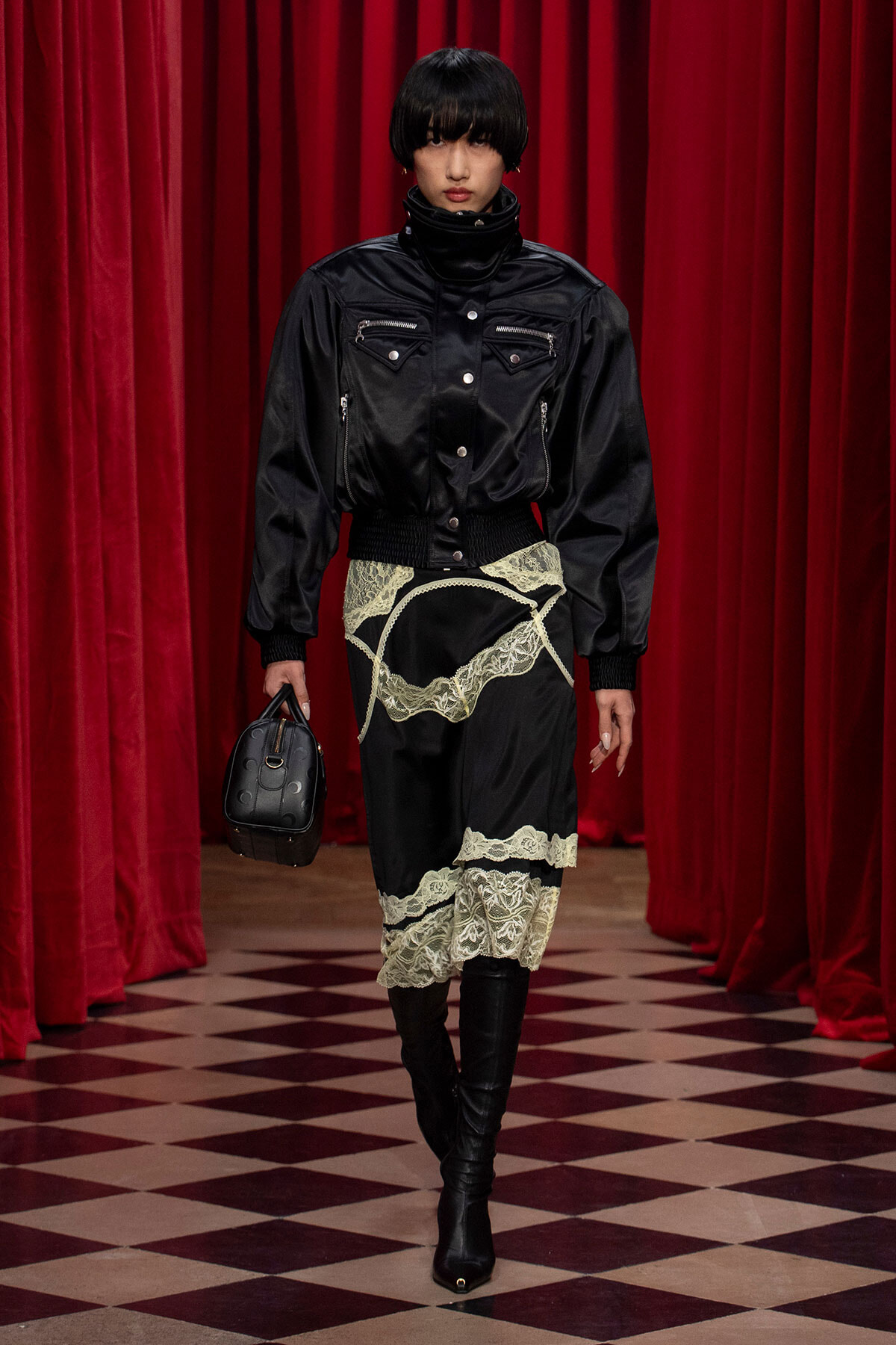 Model in black leather bomber with zip pockets, lace-overlay skirt, and knee-high boots on a red-curtain runway with a small black handbag.