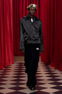 Model walking a runway in a black satin jacket, black velvet pants, white tie, and a jeweled gold crown against red curtains.