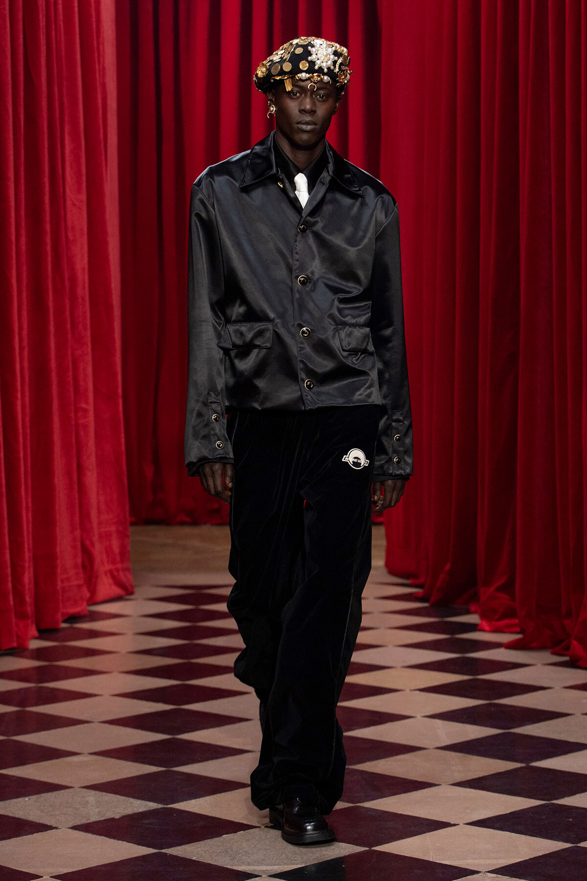 Model walking a runway in a black satin jacket, black velvet pants, white tie, and a jeweled gold crown against red curtains.
