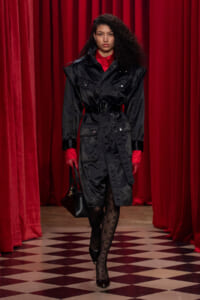 Model walking a runway in a black belted trench coat with red shirt cuffs, carrying a black handbag against red curtains and a checkered floor.
