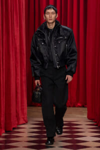 Male model in a black bomber jacket and black pants walks down a red-curtained runway, carrying a small black handbag.