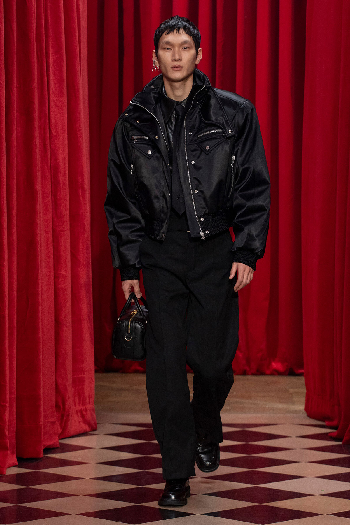 Male model in a black bomber jacket and black pants walks down a red-curtained runway, carrying a small black handbag.