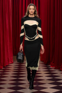 Model walking the runway in a black lace dress with cream panels, carrying a small black handbag.