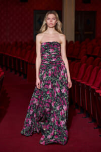 Model walks down a red theater aisle in a strapless pink and green floral maxi dress.