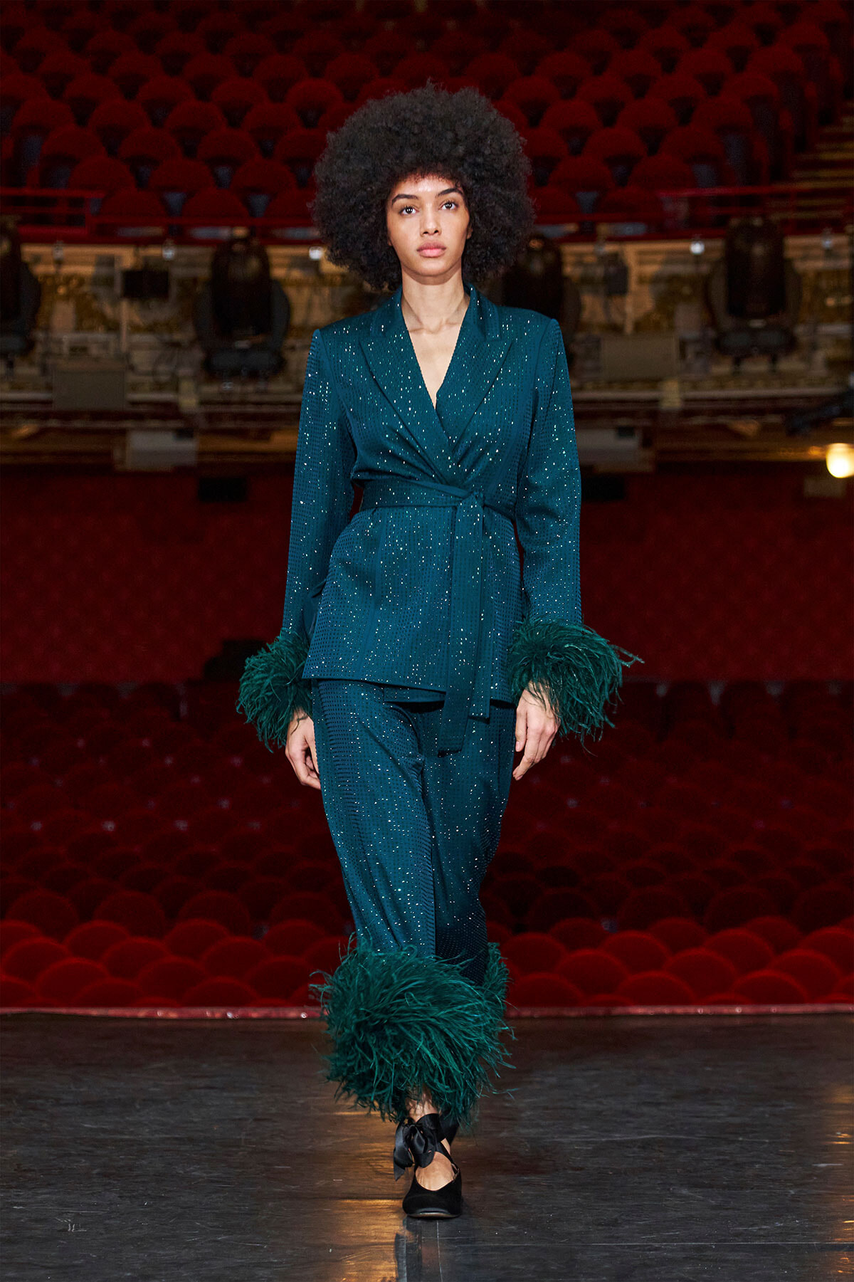 Female model walks a runway in a teal glittery blazer and matching trousers, with green feather cuffs and ankles, on a red theater set.