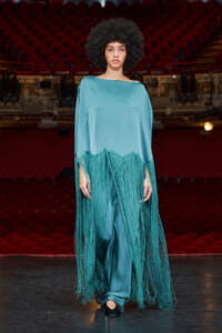 Fashion model walking on a theater stage in a teal fringe poncho over matching teal pants, dramatic long fringe swaying as she moves.