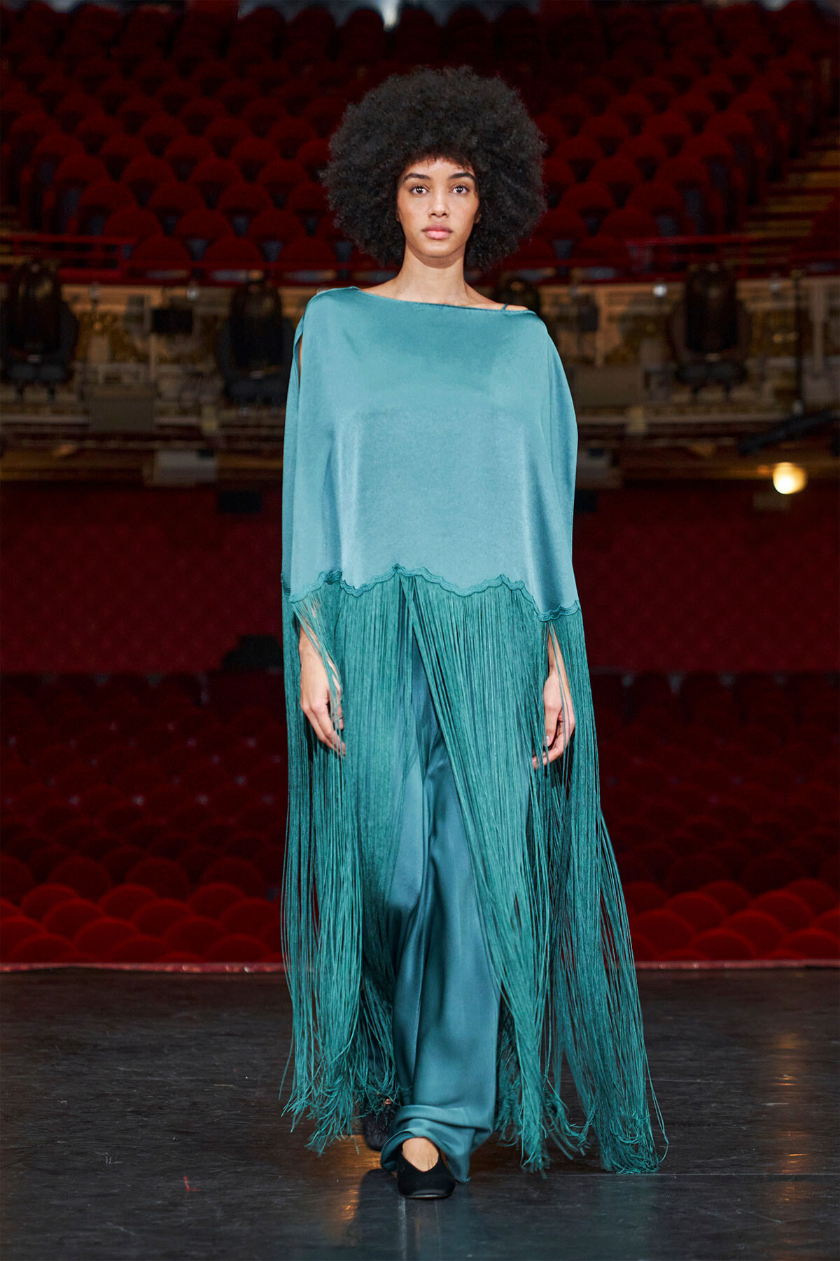 Fashion model walking on a theater stage in a teal fringe poncho over matching teal pants, dramatic long fringe swaying as she moves.