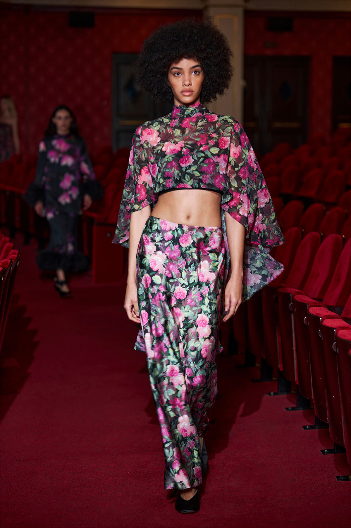 Model walks a fashion show wearing a pink floral crop top and matching wide-leg pants set on a red-carpet runway in a theater venue.