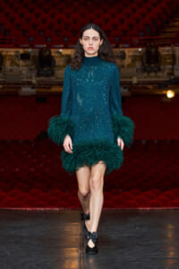 Model walking the runway in a teal sequined dress with feather trim at the sleeves and hem.