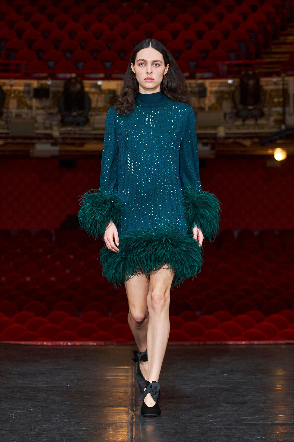Model walking the runway in a teal sequined dress with feather trim at the sleeves and hem.