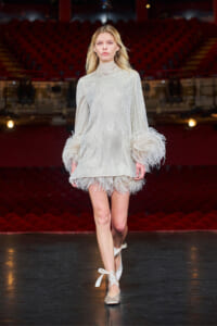 Model walks a fashion runway in a silver sequin dress with feathered cuffs and hem, theater seating in the background in red.
