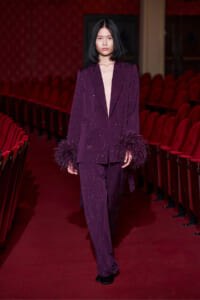 Model walks a runway in a deep purple, sequined pantsuit with feathered cuffs inside a theater with red seats.