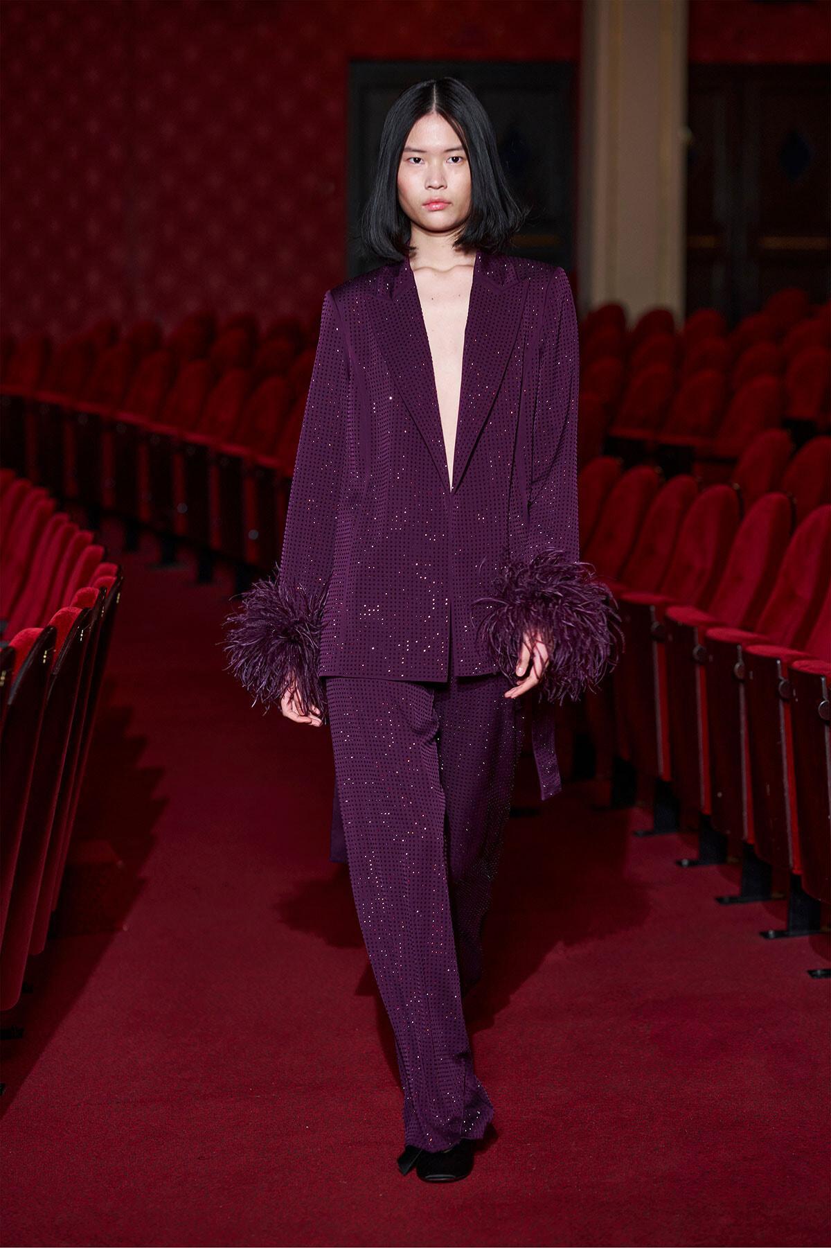 Model walks a runway in a deep purple, sequined pantsuit with feathered cuffs inside a theater with red seats.