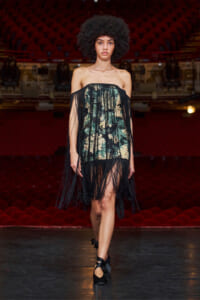 Model walking a runway in a strapless floral dress with long black fringe overlay, backstage with red theater seats in view.