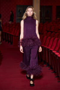 Model walks a purple sequined sleeveless jumpsuit with feather trims at wrists and hems along a red theater runway.