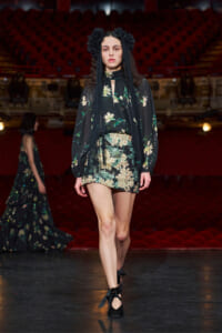 Model walks a runway in a black floral mini dress with long sheer sleeves and a black flower headpiece, on a dimly lit stage with red seats behind her.