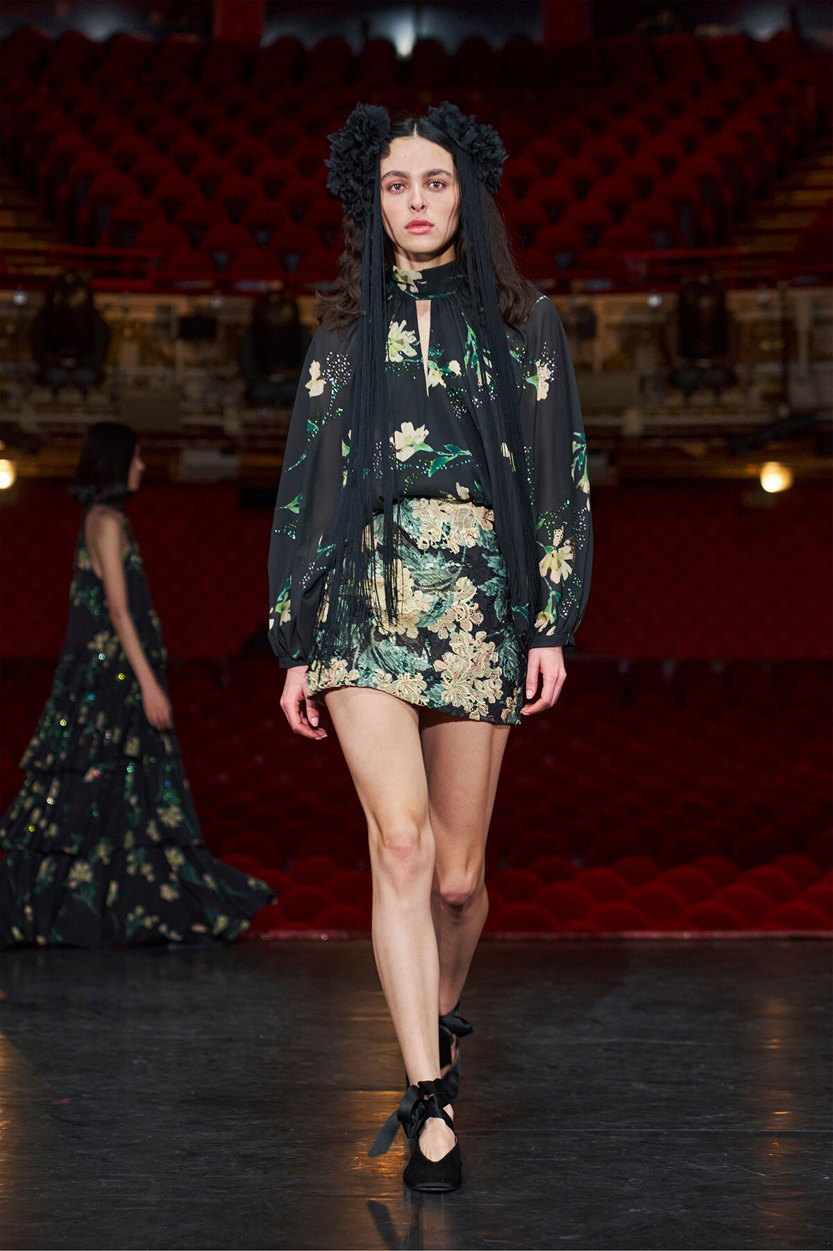 Model walks a runway in a black floral mini dress with long sheer sleeves and a black flower headpiece, on a dimly lit stage with red seats behind her.