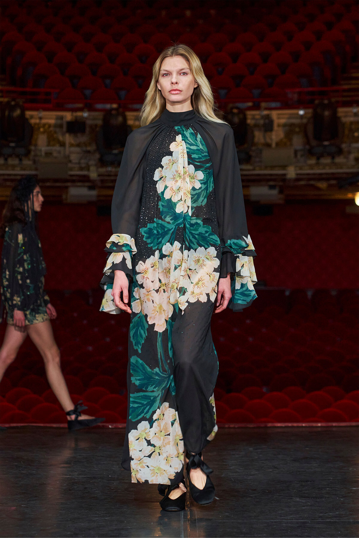 Female model wearing a long black floral dress with cape sleeves on a fashion runway.
