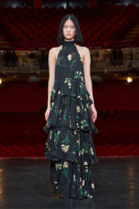 Model wearing a black floral tiered gown on a stage with red theater seats in the background.