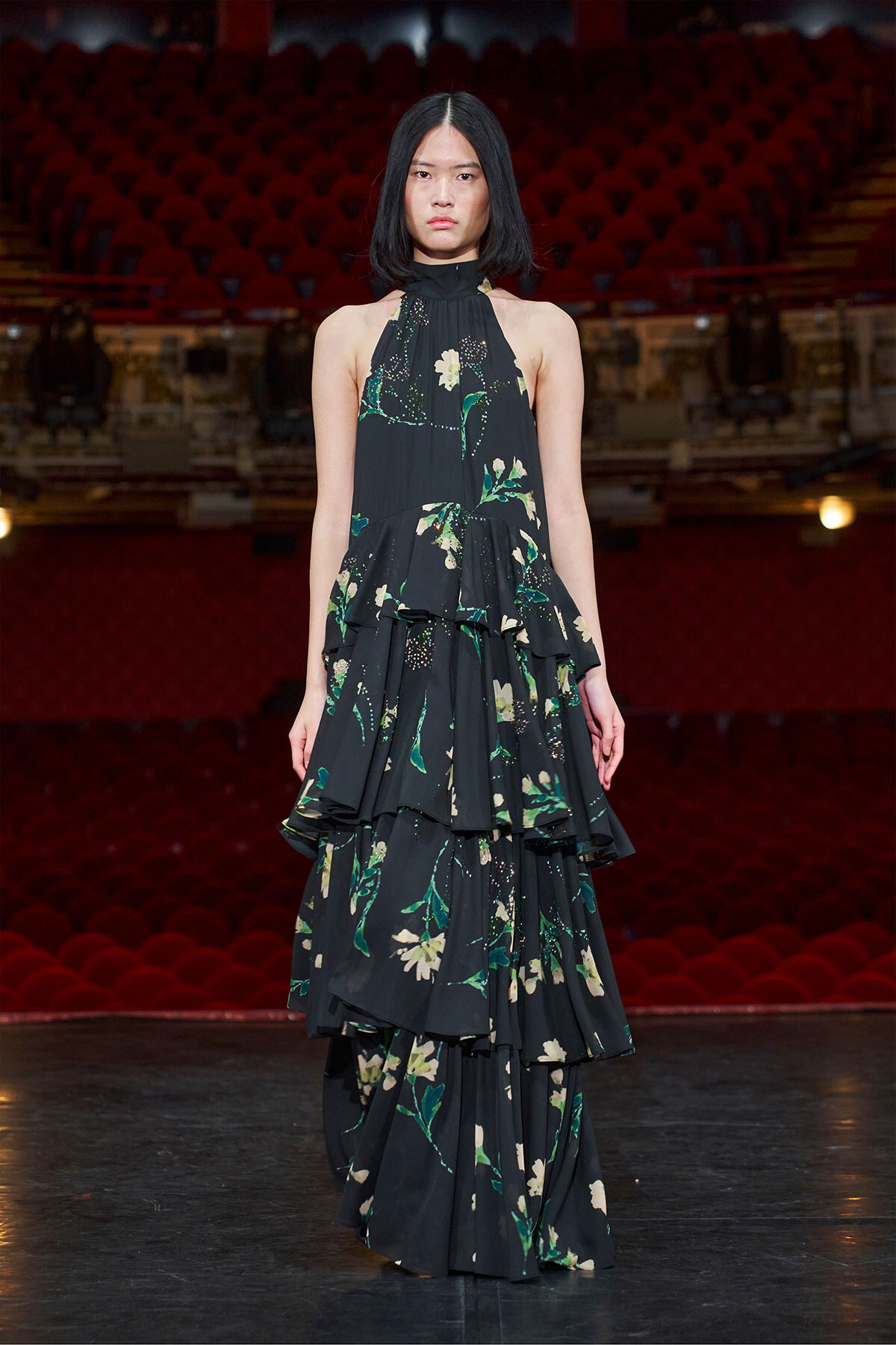 Model wearing a black floral tiered gown on a stage with red theater seats in the background.