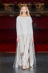 Model walking a white fringe dress on a runway in a theater with red seats in the background.