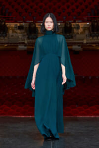 Full-length teal gown with a high collar and cape-like sleeves, modeled on a stage in a theater with red seats in the background.