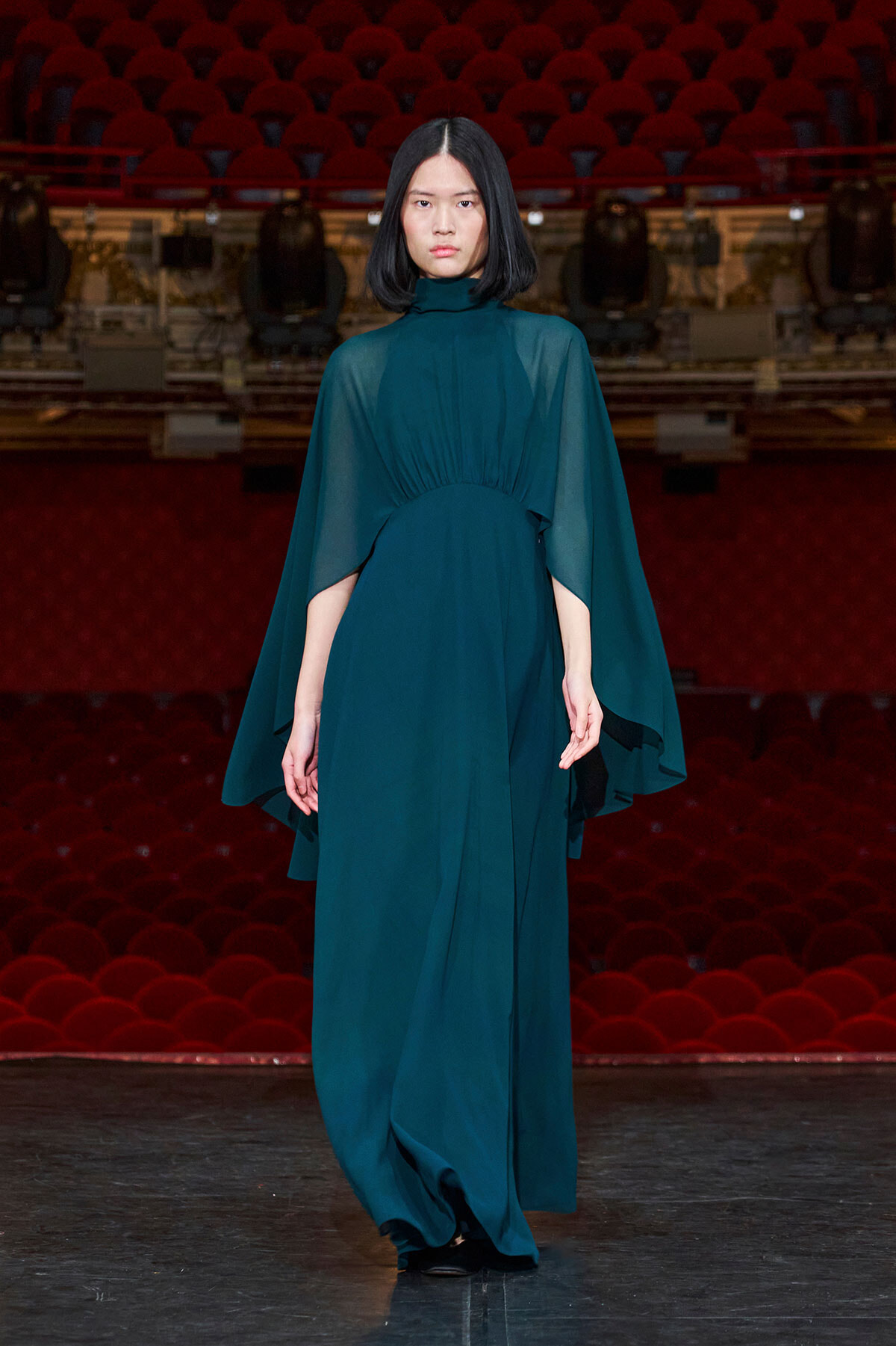 Full-length teal gown with a high collar and cape-like sleeves, modeled on a stage in a theater with red seats in the background.