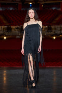 Model walking a runway in a black fringe dress with spaghetti straps and long fringe details.