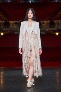 Female model walking a fashion show runway in a pale blazer with a lace-fringe overlay, white dress underneath, and satin bow-tied flats; red theater seating in the background