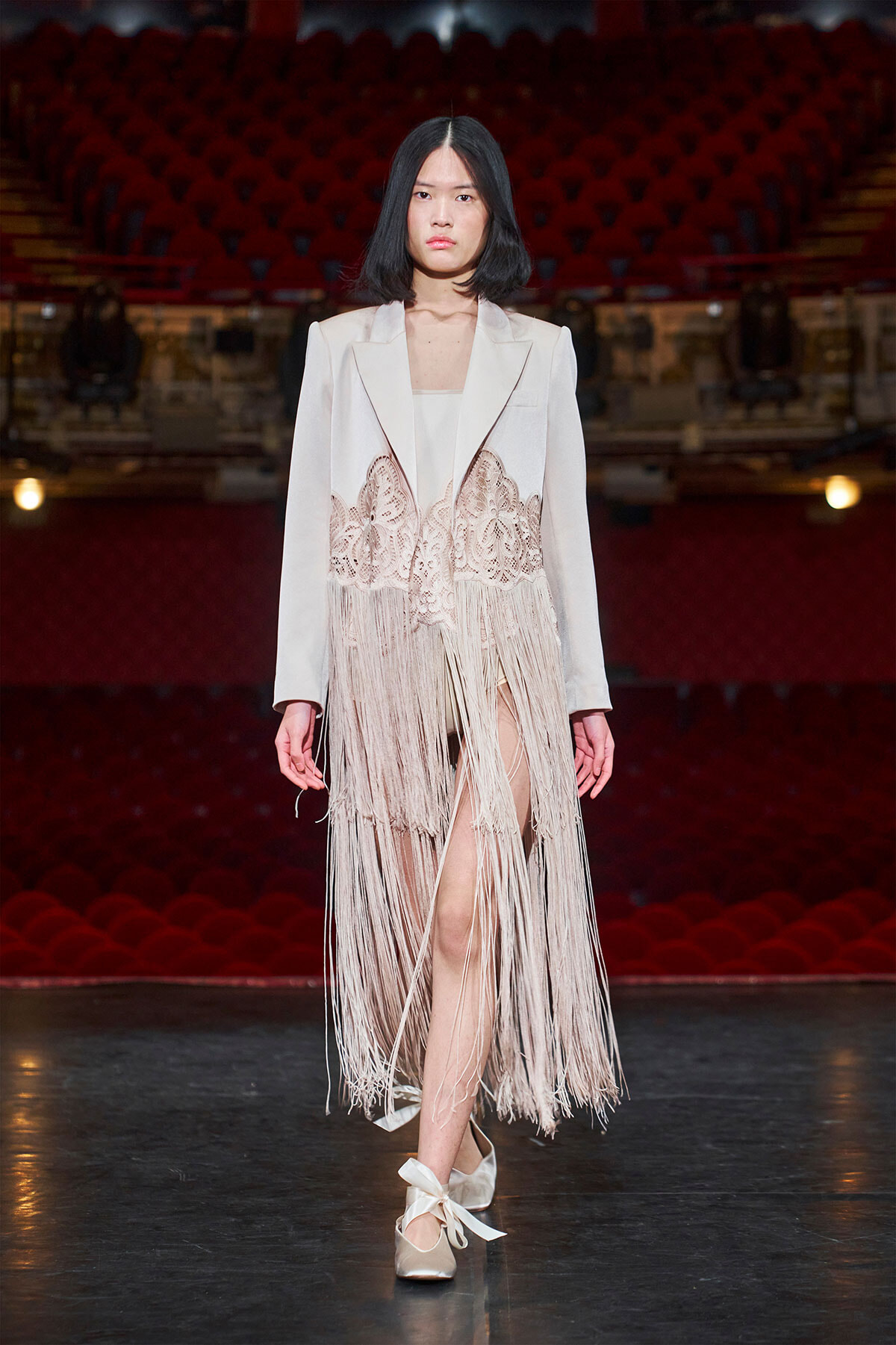 Female model walking a fashion show runway in a pale blazer with a lace-fringe overlay, white dress underneath, and satin bow-tied flats; red theater seating in the background