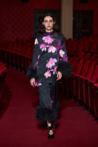 Model walking a runway in a dark floral tunic and pants with black feather trim at cuffs and ankles in a red-velvet theater setting.