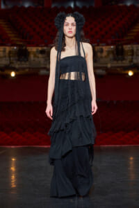 Model walking a fashion runway in a black fringe two-piece outfit with long fringe strands and a floral headpiece in a red theater setting