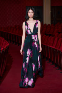 Model in a deep green floral gown walking down a red-carpet runway surrounded by red theater seats.