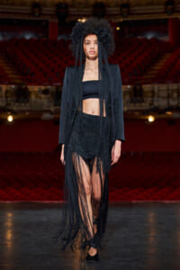 Model walks the runway in a black ensemble with long fringe details, including a cropped top, blazer, and a floral headpiece.
