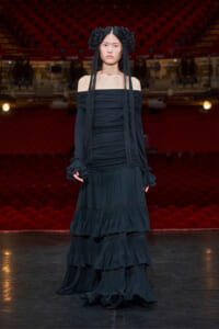 Model on a theater stage wearing a long black off-the-shoulder dress with tiered ruffles and large floral headpieces in her hair.