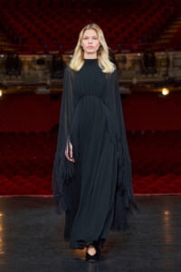 Model walks a runway in a long black dress with sheer sleeves and heavy fringe at the hem, on a stage with red theater seating behind.