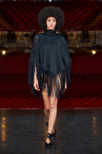 Model walking the runway in a black fringed poncho dress with lace sleeves and black heels, theater seating in the background.